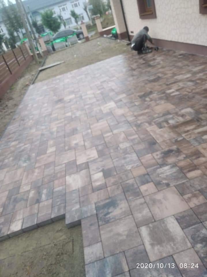 Układanie kostki brukowej w odcieniach szarości i brązu, widoczny fragment niedokończonej powierzchni i pracownik w tle, dom z jasną elewacją i brązowymi oknami.