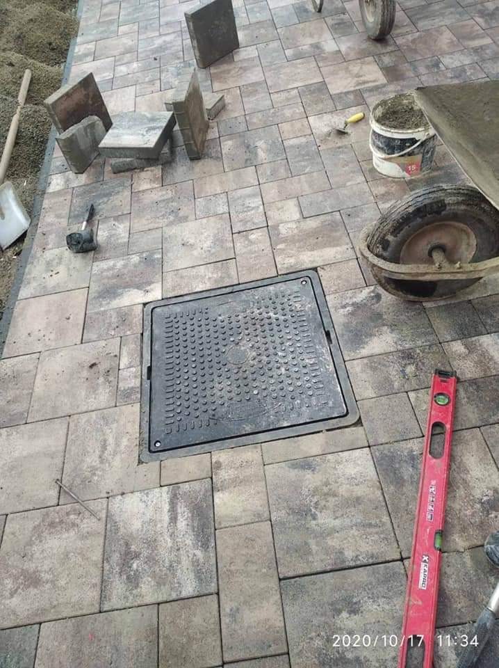 Ułożona kostka brukowa w odcieniach szarości z włazem kanalizacyjnym pośrodku, widoczne narzędzia brukarskie: poziomica, młotek, taczka i wiadro z zaprawą.