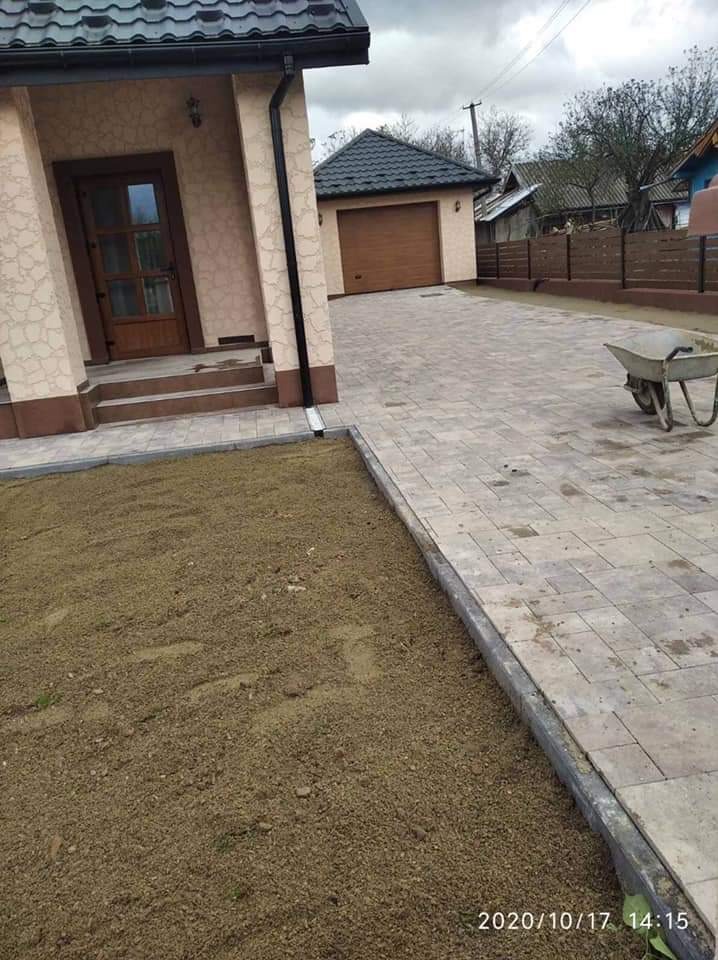 Ułożona szara kostka brukowa na podjeździe z widocznym domem i garażem w tle, obok przygotowany teren pod trawnik, taczka stoi na boku.