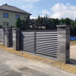 Nowoczesna brama wjazdowa przesuwna z poziomymi panelami, osadzona między betonowymi słupkami, widoczna z ulicy przed domem z basenem w tle.