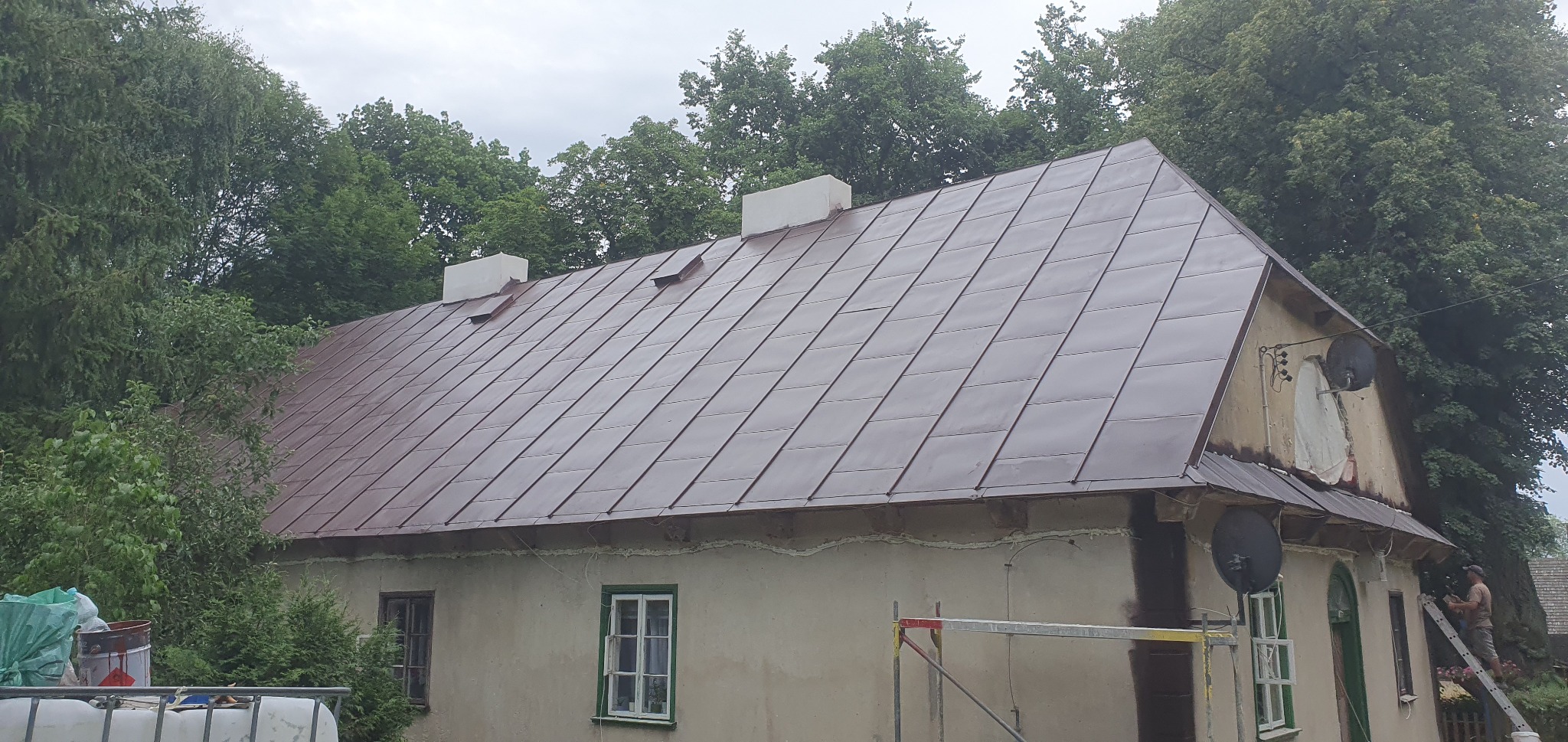 Odnowiony, brązowy dach z blachy na starym domu z dwoma kominami, widoczny pracownik na drabinie podczas prac wykończeniowych.