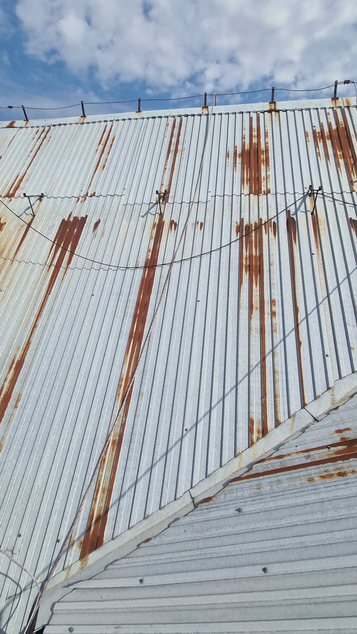 Skorodowany blaszany dach z widocznymi śladami rdzy i systemem odgromowym, sfotografowany z niskiego kąta, ukazujący fragment nieba z chmurami.