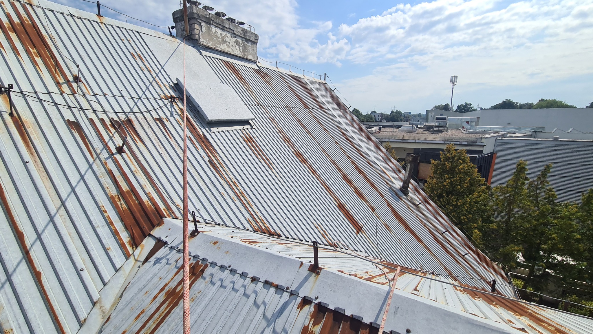 Metalowy dach z widocznymi śladami rdzy i mocowaniami lin asekuracyjnych, na tle błękitnego nieba z chmurami.