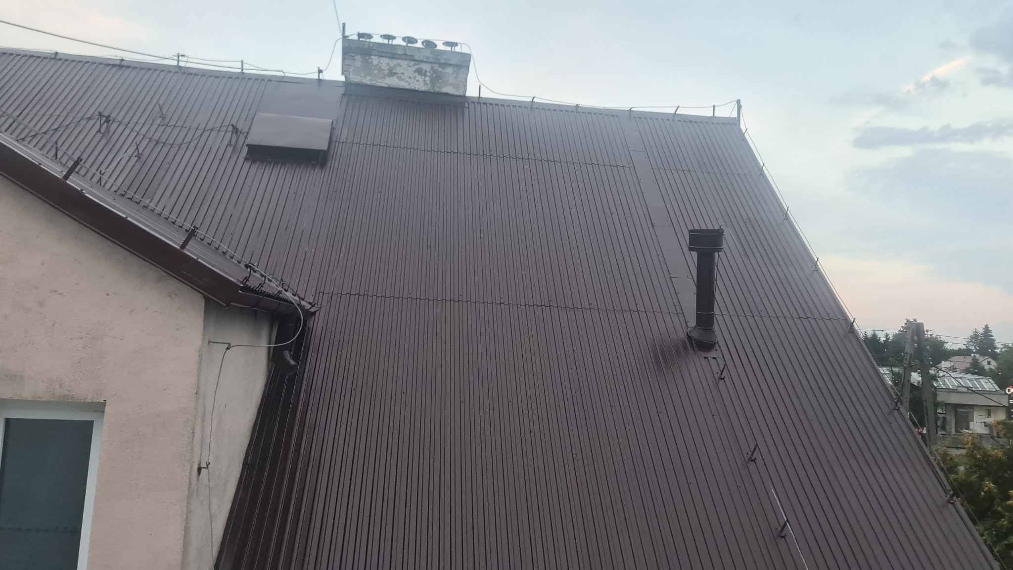 Widok z góry na brązowy, blaszany dach domu jednorodzinnego z widocznym kominem, wentylacją i rynnami przy ścianie budynku.