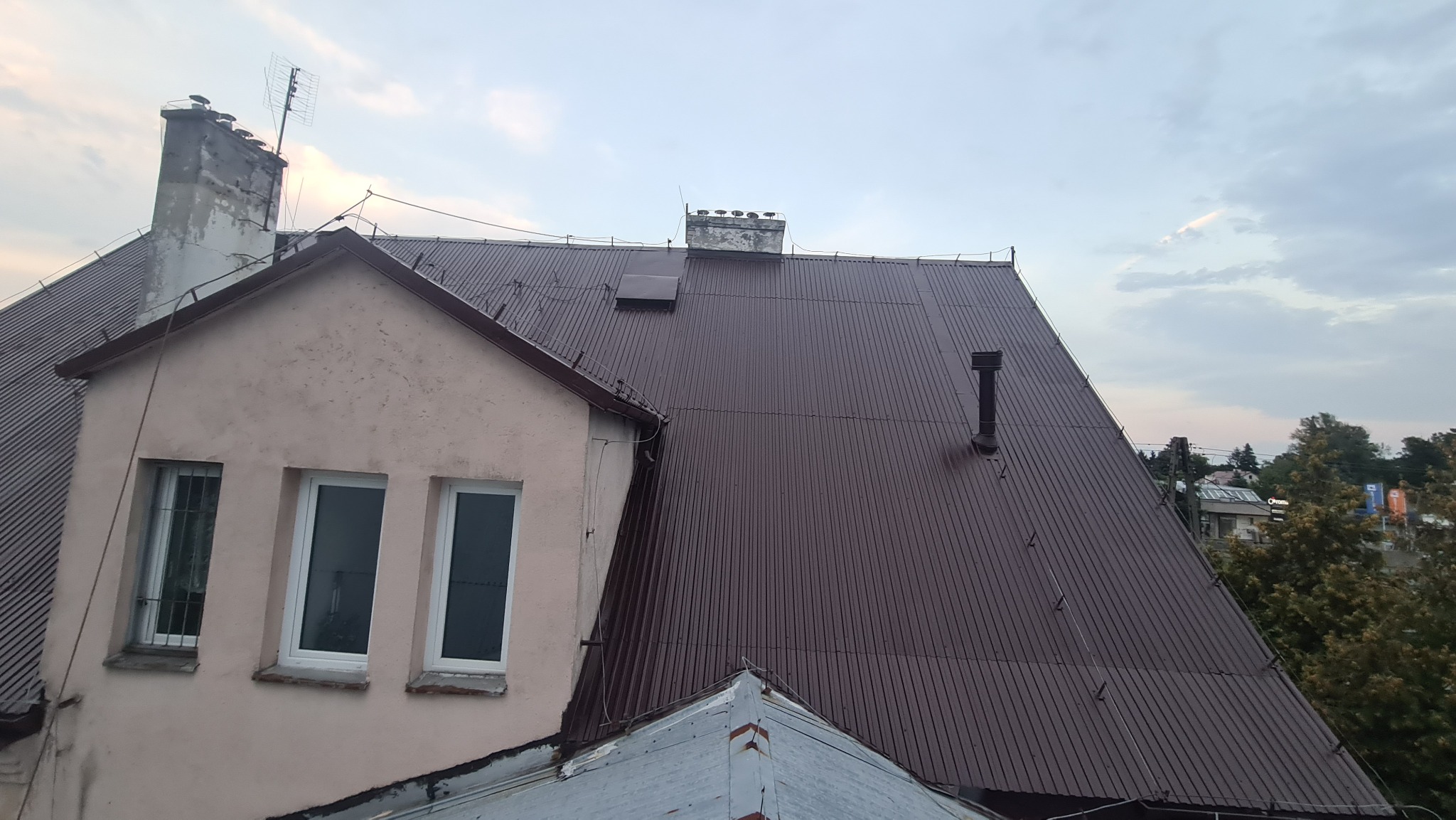 Ciemnobrązowy blaszany dach z kominem i anteną telewizyjną na tle pochmurnego nieba, widok z góry.