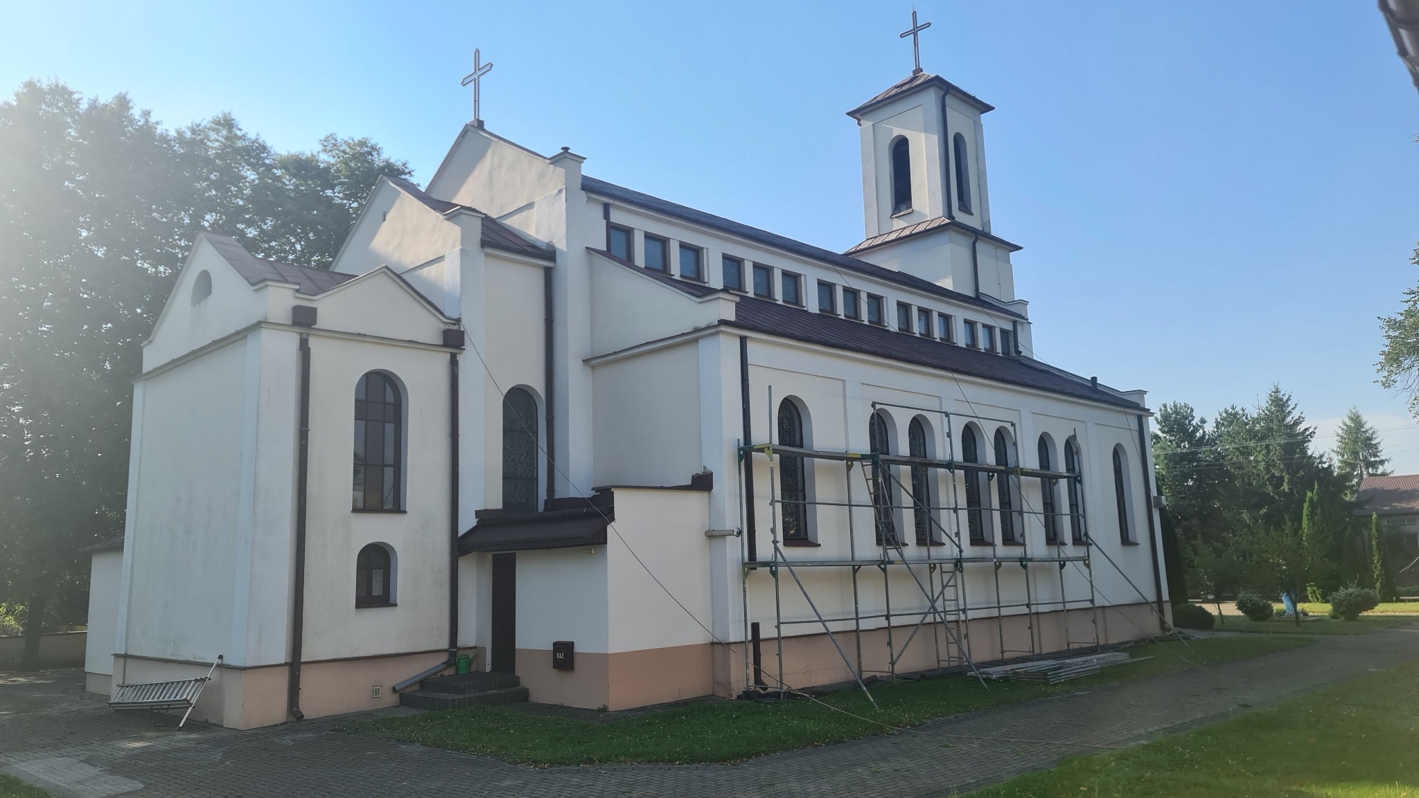 Kościół z brązowym dachem, białą elewacją i rusztowaniem przy bocznej ścianie, widoczne drzewa w tle.