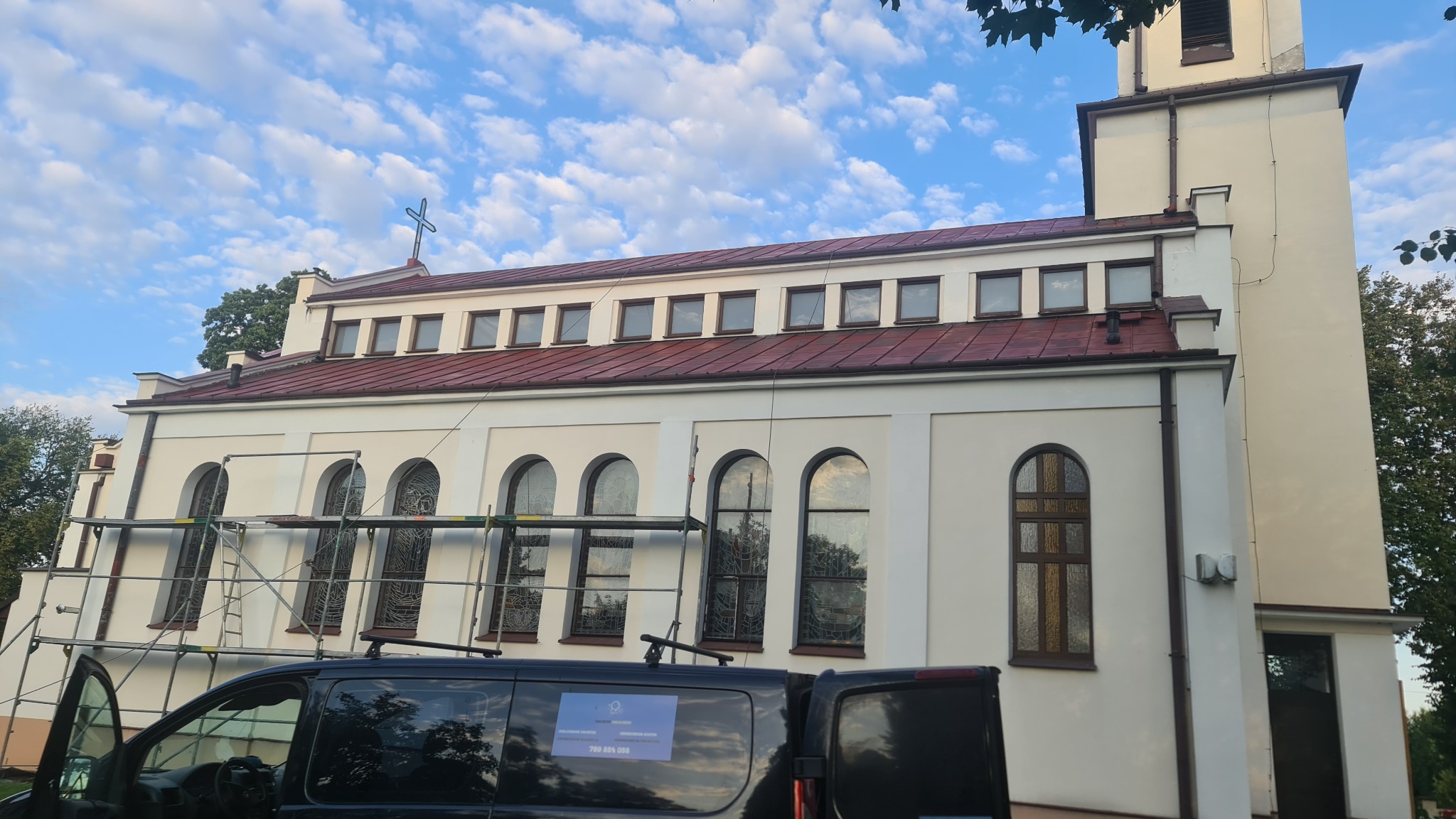 Odrestaurowany dach kościoła z krzyżem, widoczny z rusztowania i samochodu firmy budowlanej w słoneczny dzień.