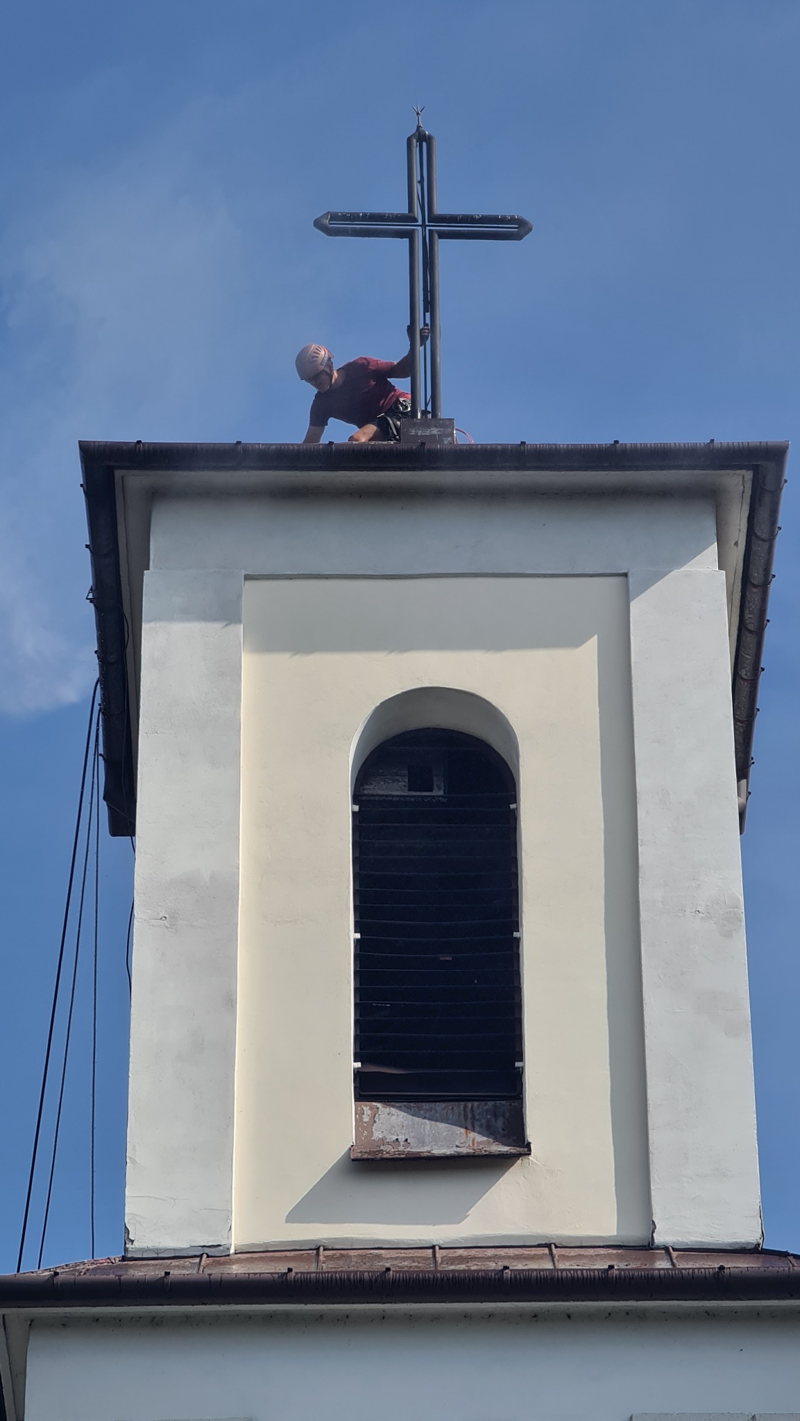 Pracownik w kasku na dachu wieży kościelnej, przy metalowym krzyżu, prawdopodobnie podczas prac konserwacyjnych.