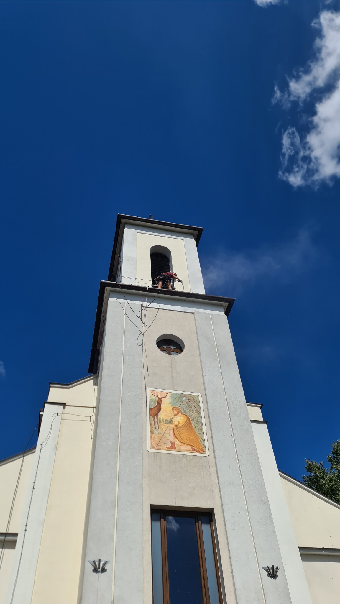 Pracownik zabezpieczony linami na wysokości, podczas prac konserwatorskich na dachu wieży kościelnej z malowidłem religijnym pod okrągłym oknem.