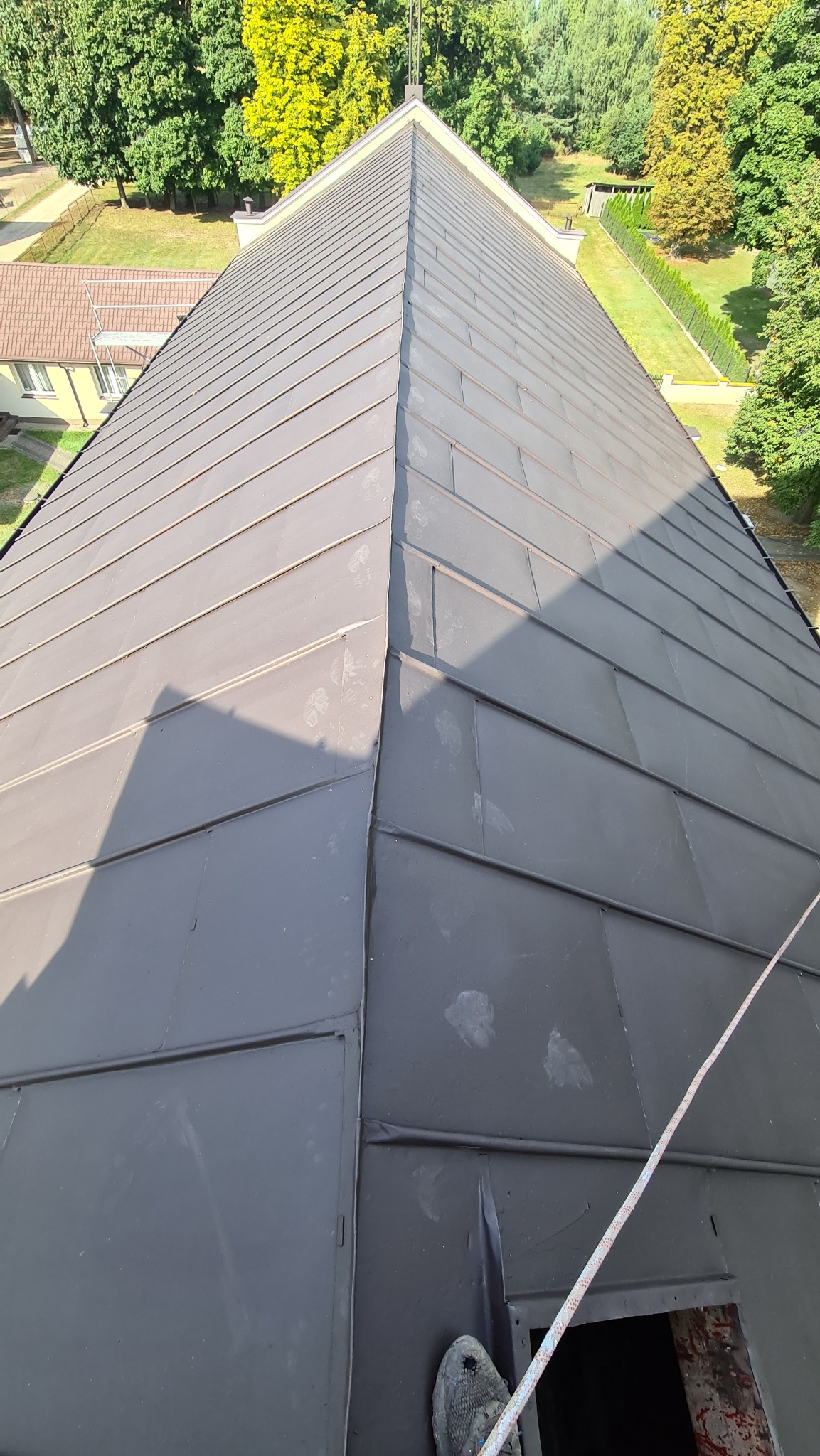 Widok z góry na pomalowany na szaro dach z blachy z widocznymi śladami stóp i liną zabezpieczającą, w tle zieleń drzew i zabudowa.