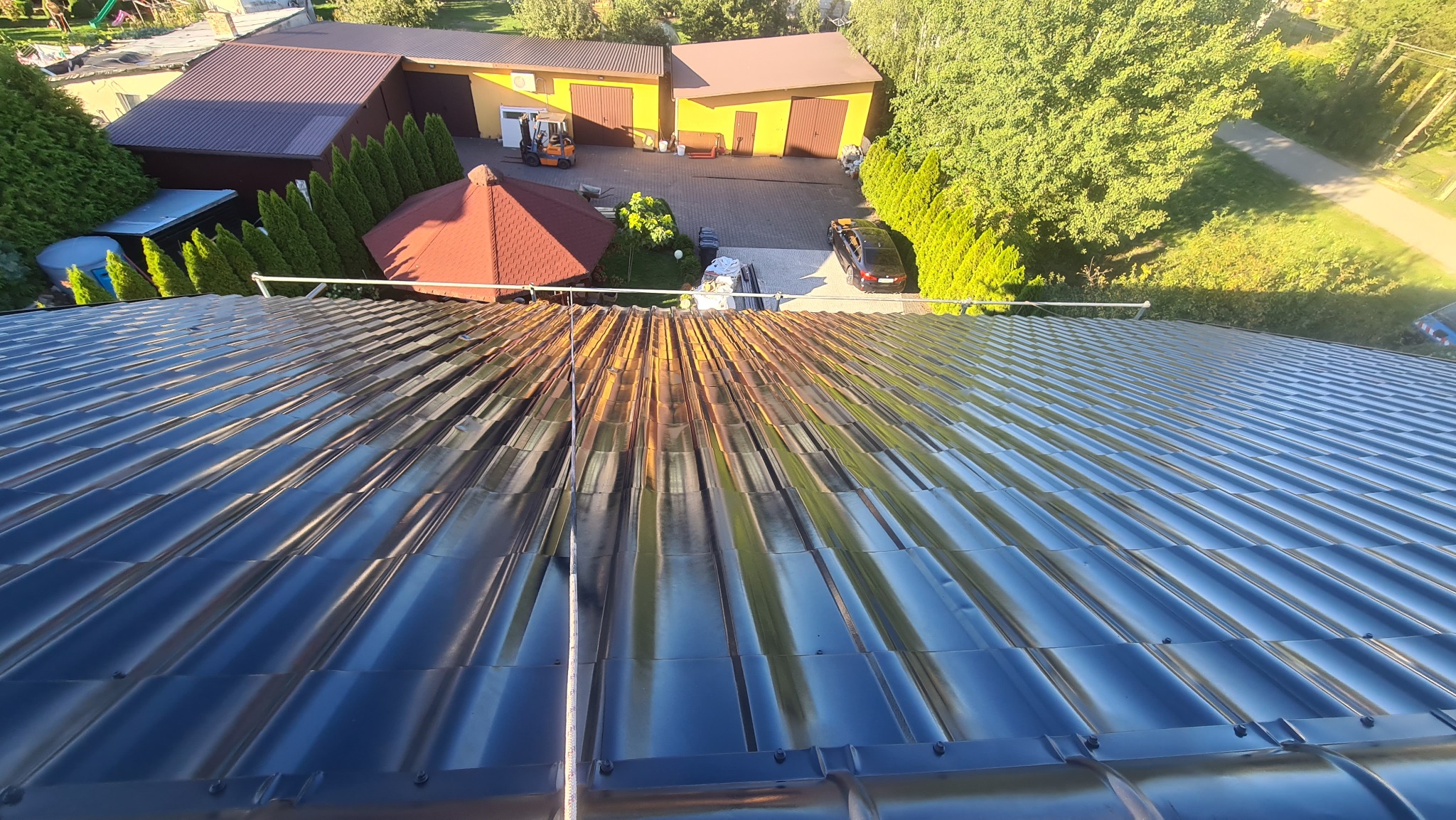Widok z góry na metalowy dach z widocznymi śladami korozji i przebarwień, w tle budynki gospodarcze i zieleń.