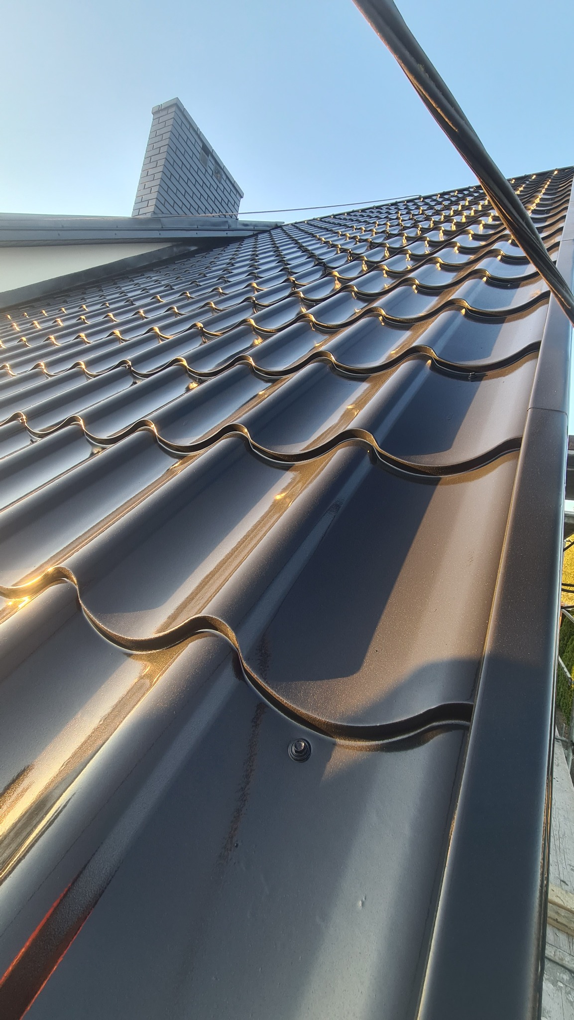 Błyszczące, ciemne dachówki ceramiczne na dachu domu w promieniach zachodzącego słońca, widoczny komin z szarej cegły i fragment rynny.