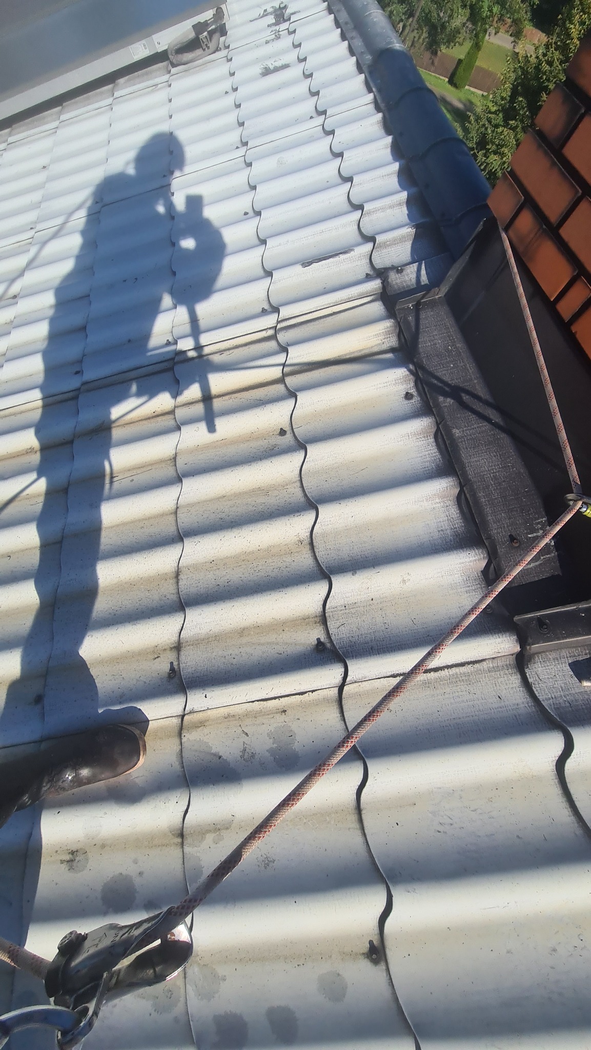 Biały dach z blachy falistej, cień osoby z telefonem, lina zabezpieczająca przymocowana do dachu.