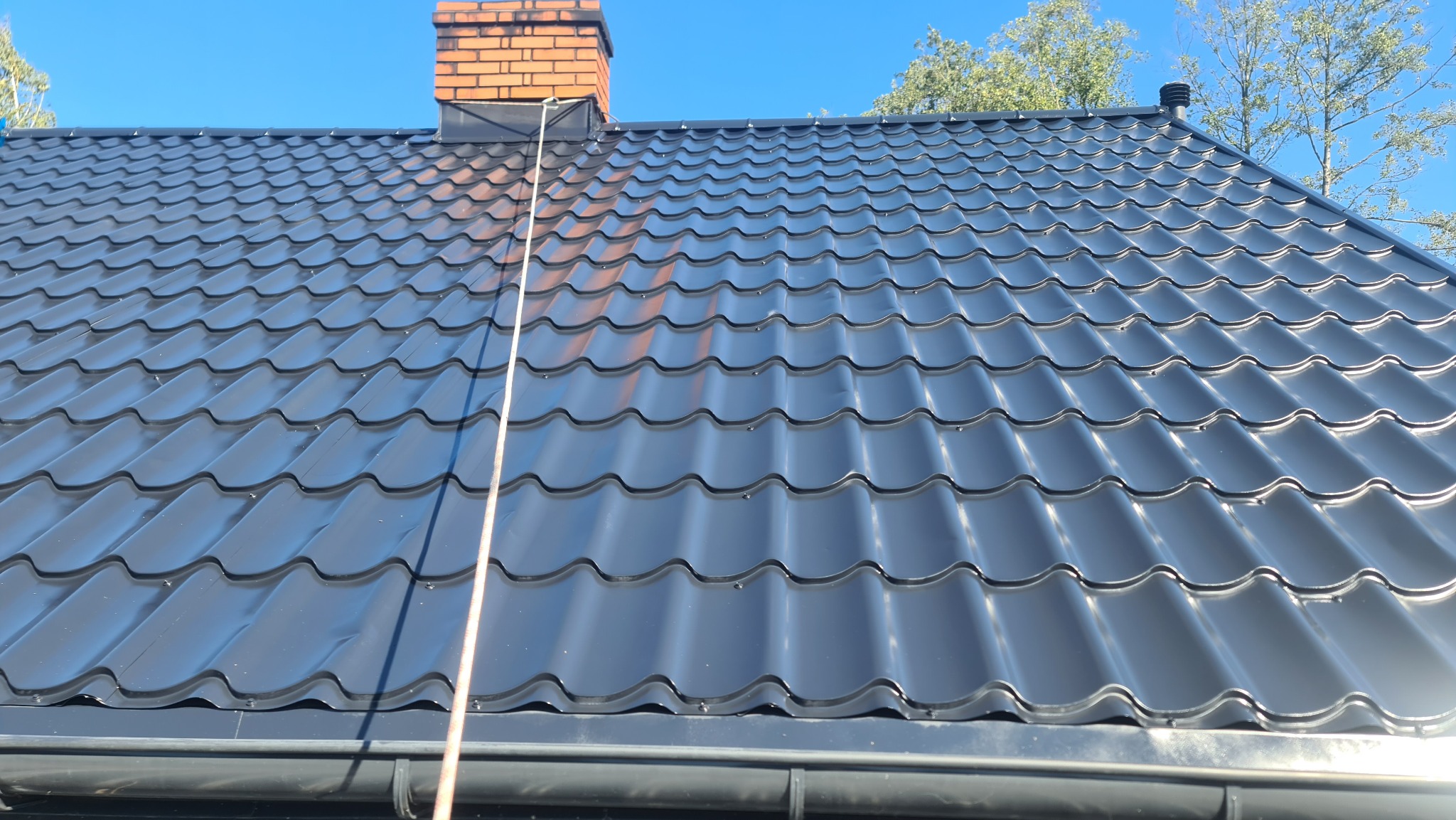 Błyszczący, nowy, grafitowy dach z metalowej dachówki modułowej, widoczny ceglany komin i czysta rynna w słoneczny dzień.
