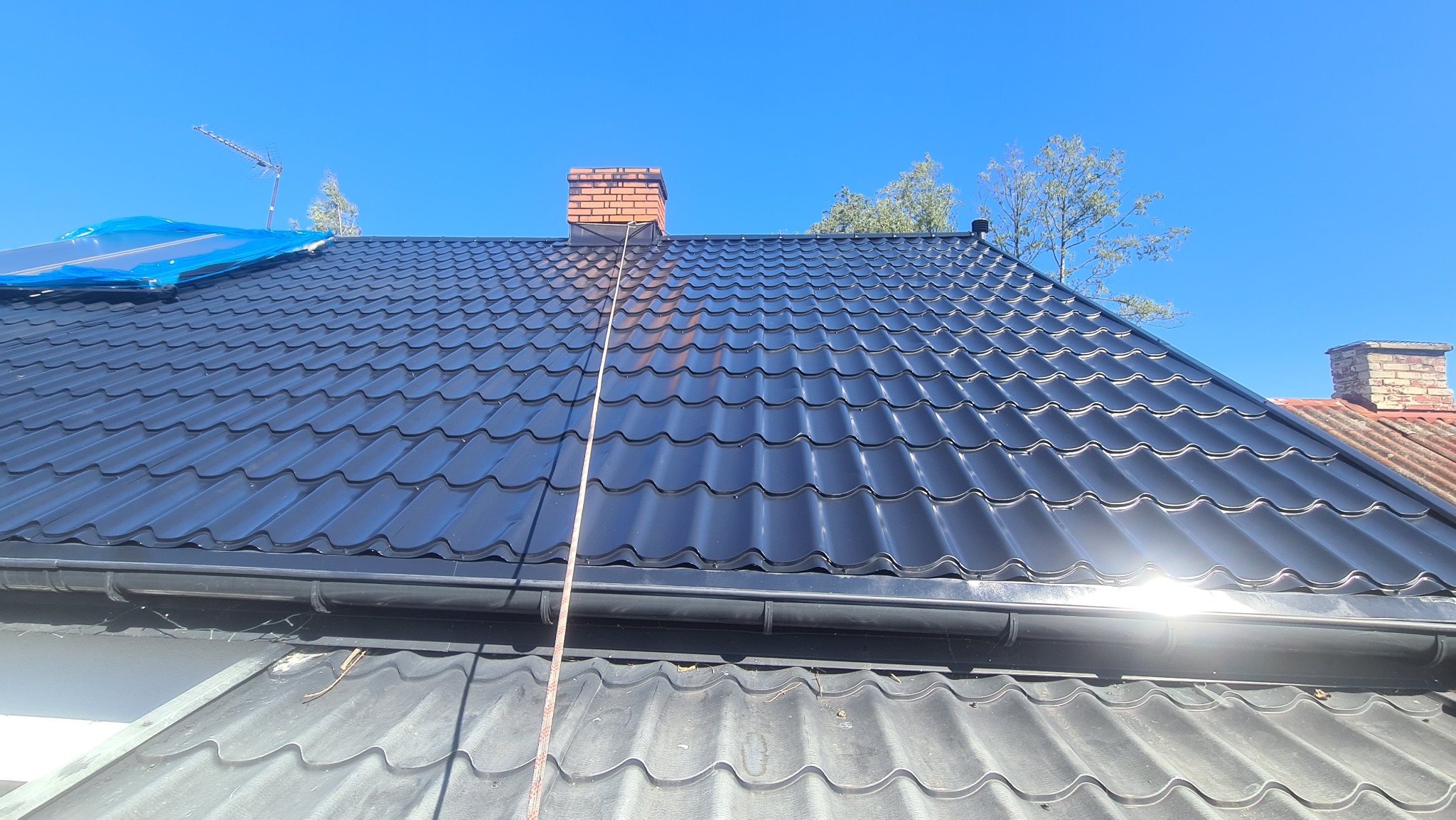 Błyszczący, czarny dach z blachodachówki po malowaniu w Tłuszczu, widok na komin z cegły i rynnę, część dachu zabezpieczona niebieską folią.