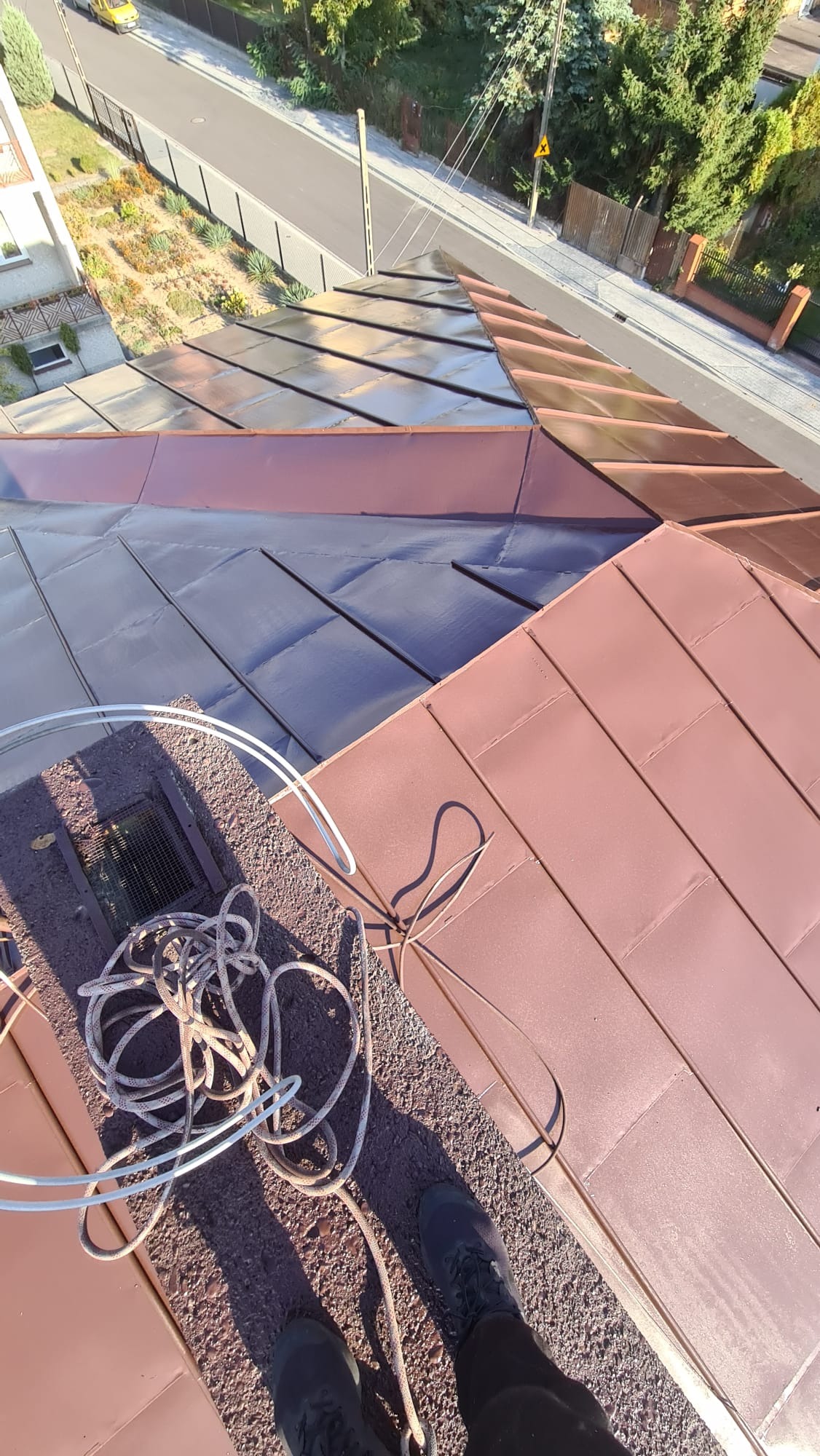 Widok z góry na dach z blachy trapezowej pomalowany na kolor Ral 8017, częściowo pomalowany na ciemniejszy odcień, na dachu leży zwinięta lina zabezpieczająca, w tle widoczna ulica i zabudowania.