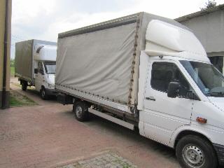 Dwa białe busy z plandekami zaparkowane na brukowanej drodze, widoczne z boku, z budynkiem w tle. Jeden bus częściowo zasłania drugi.