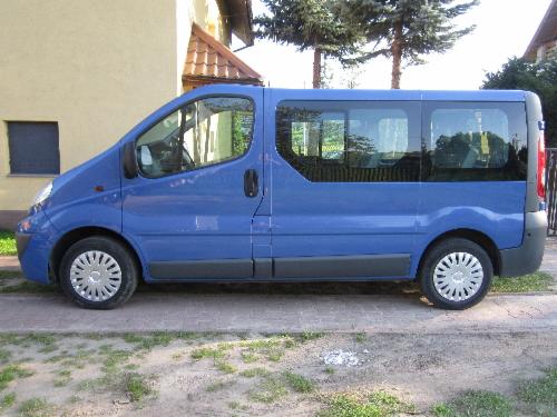 Niebieski van z przyciemnianymi szybami, zaparkowany przed budynkiem. Widok z boku, oświetlenie dzienne. Koła z srebrnymi felgami.