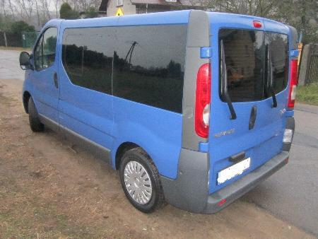 Niebieski bus Renault Trafic z przyciemnianymi szybami, zaparkowany na poboczu drogi, widziany z tyłu pod kątem. Widoczne światła stop i wycieraczki.