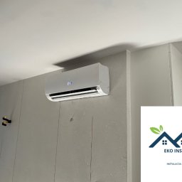EKO INS-TECH - Klimatyzator ścienny z wyświetlaczem ustawionym na 21 stopni Celsjusza, zamontowany na szarej ścianie z pionowymi panelami dekoracyjnymi, widoczne logo firmy EKO INS-TECH w prawym dolnym rogu.