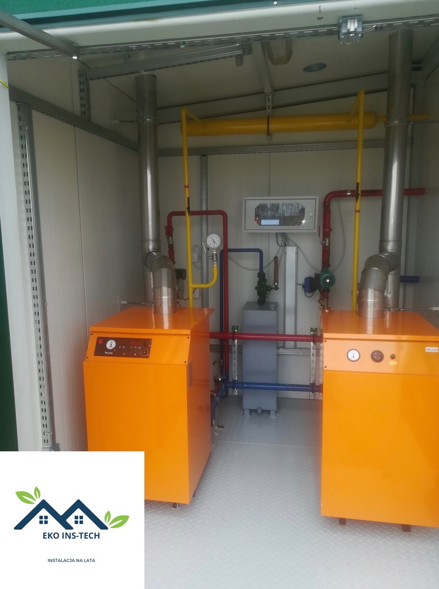 Dwa pomarańczowe piece centralnego ogrzewania w kontenerze, z widocznymi rurami instalacji w kolorach żółtym, czerwonym i niebieskim, panel sterowania na ścianie i logo firmy Eko Ins-Tech w lewym...
