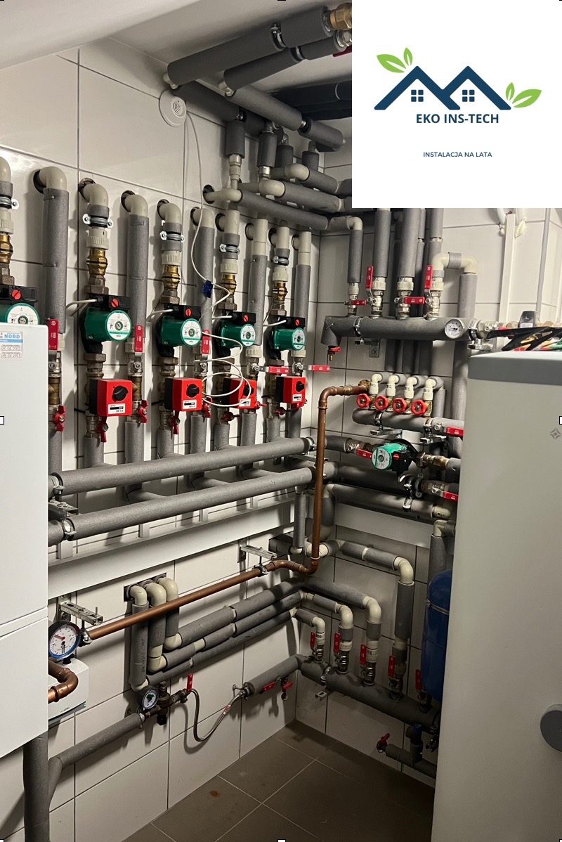 Kompleksowa instalacja grzewcza z licznymi rurami, zaworami, pompami obiegowymi i izolacją termiczną, zamontowana na ścianie wyłożonej białymi płytkami, widoczny fragment zasobnika ciepłej wody...