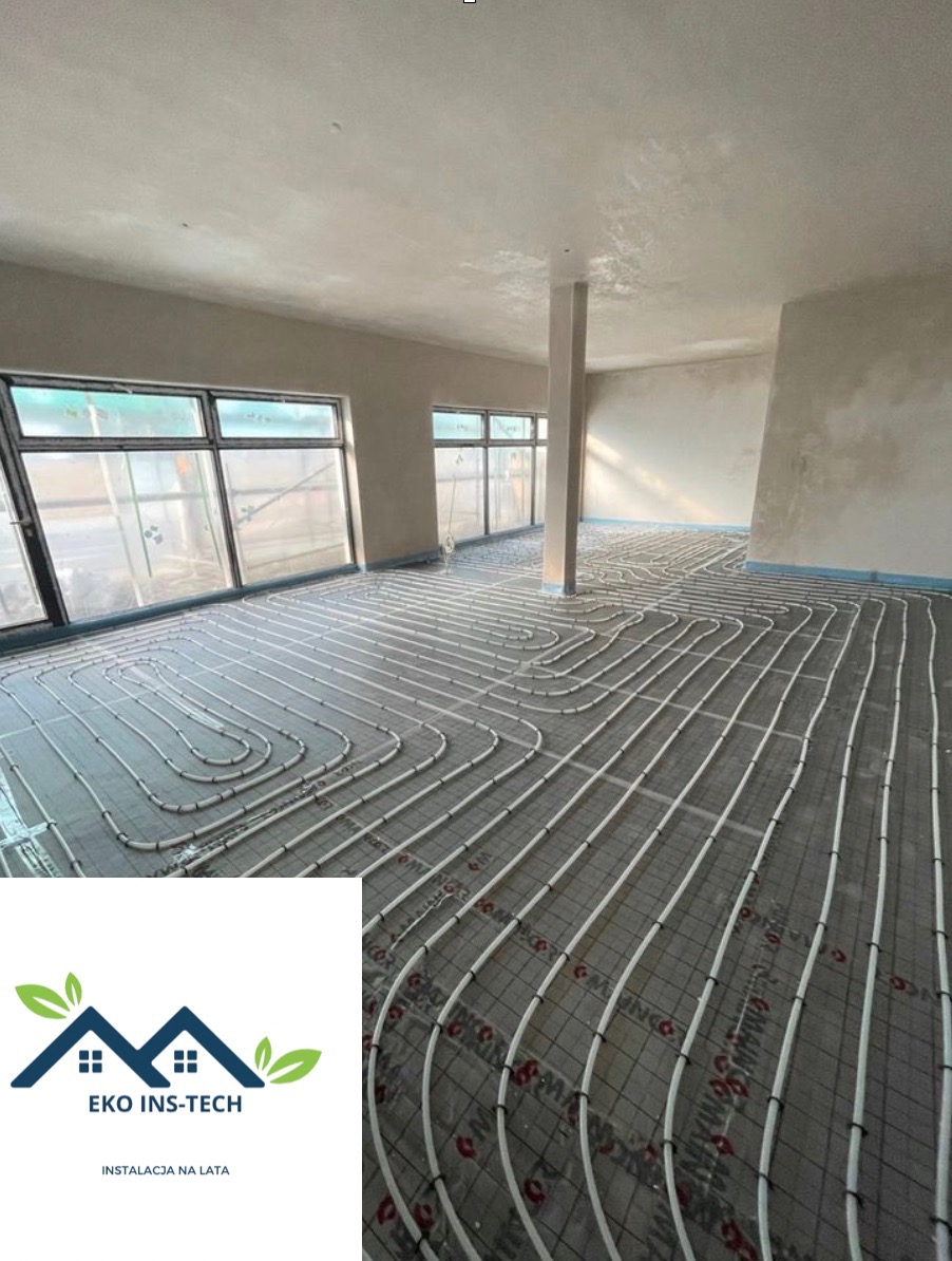 Rozłożona instalacja ogrzewania podłogowego w surowym pomieszczeniu z dużymi oknami, widoczny betonowy podkład i białe rurki mocowane do siatki, w rogu logo firmy EKO INS-TECH.