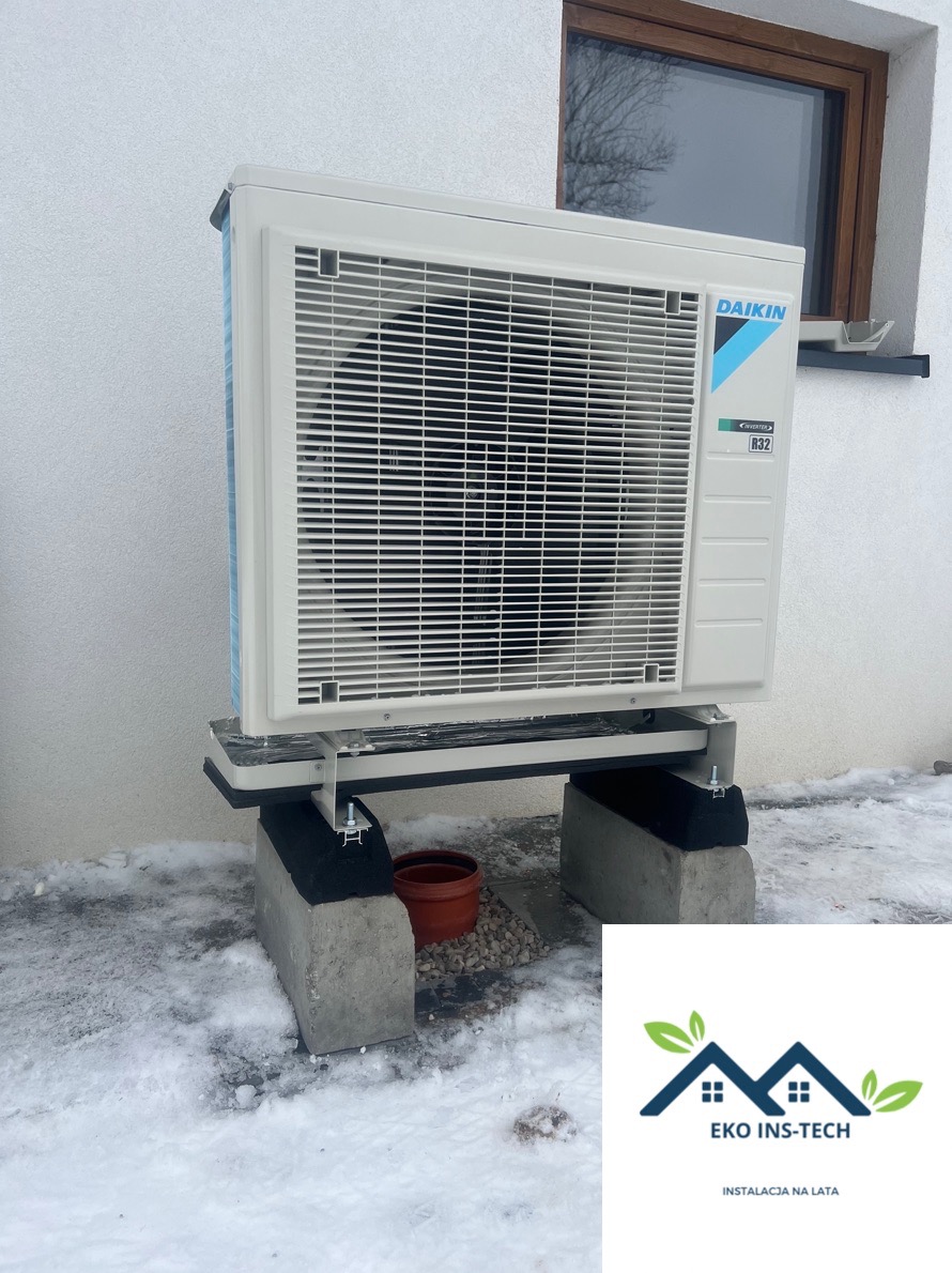 Biała zewnętrzna jednostka pompy ciepła Daikin na betonowych blokach w zimowym otoczeniu, z widocznym oknem w tle i logiem firmy Eko Ins-Tech nałożonym na zdjęcie.