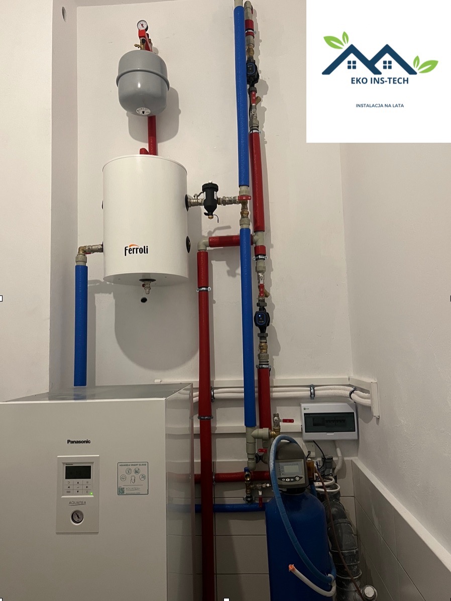 Zainstalowany biały piec Ferroli z szarym zbiornikiem wyrównawczym, połączony z instalacją z rur czerwonych i niebieskich, obok biała pompa ciepła Panasonic Aquarea, widoczny też filtr wody...