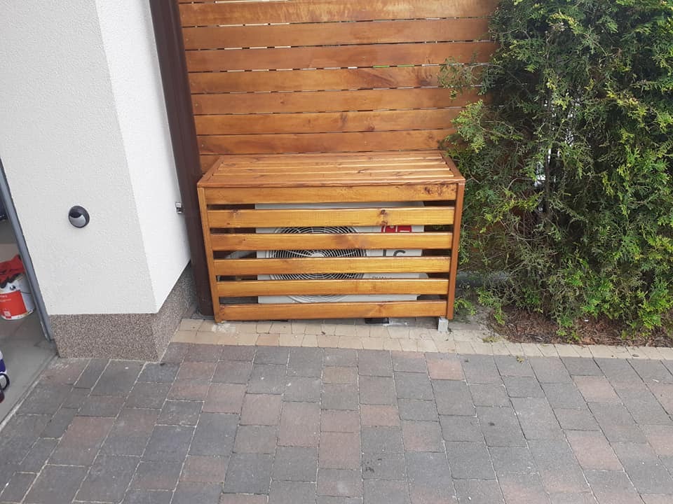 Zewnętrzna jednostka klimatyzacji ukryta w drewnianej obudowie z poziomych listew, ustawiona przy ścianie domu obok drewnianego ogrodzenia i krzewu.