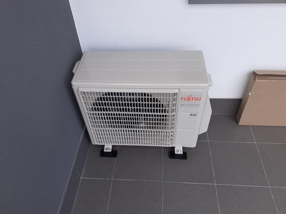 Kompaktowa jednostka zewnętrzna klimatyzatora Fujitsu DC Inverter R32 ustawiona na szarych płytkach pod ścianą pomalowaną na dwa kolory.