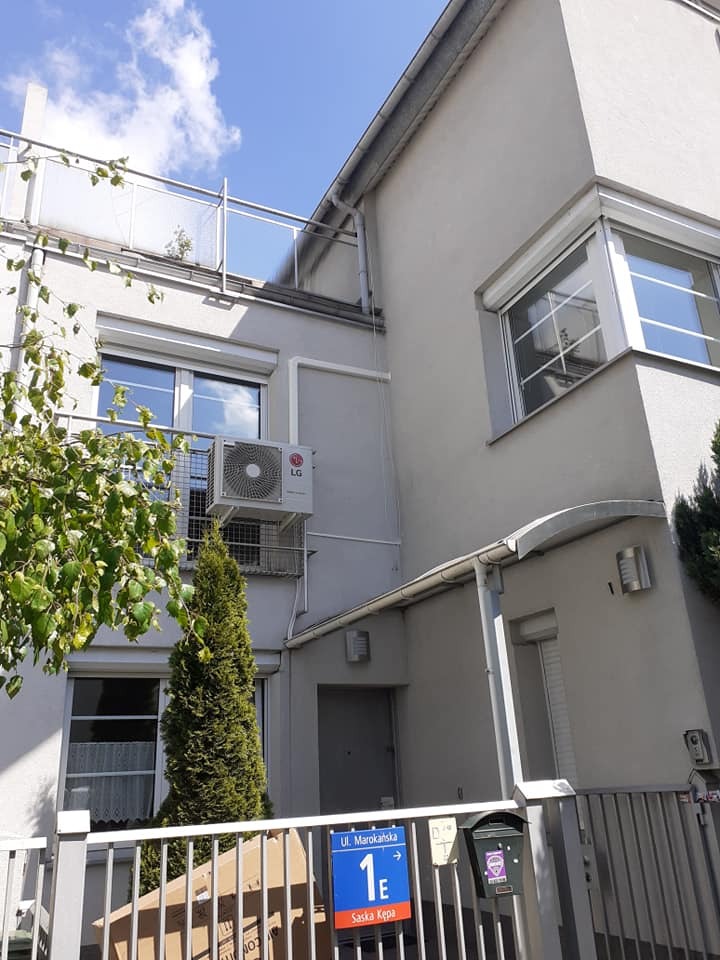 Zewnętrzna jednostka klimatyzacji LG zamontowana na ścianie budynku mieszkalnego z balkonem, widok z poziomu ulicy Marokańskiej na Saskiej Kępie w słoneczny dzień.