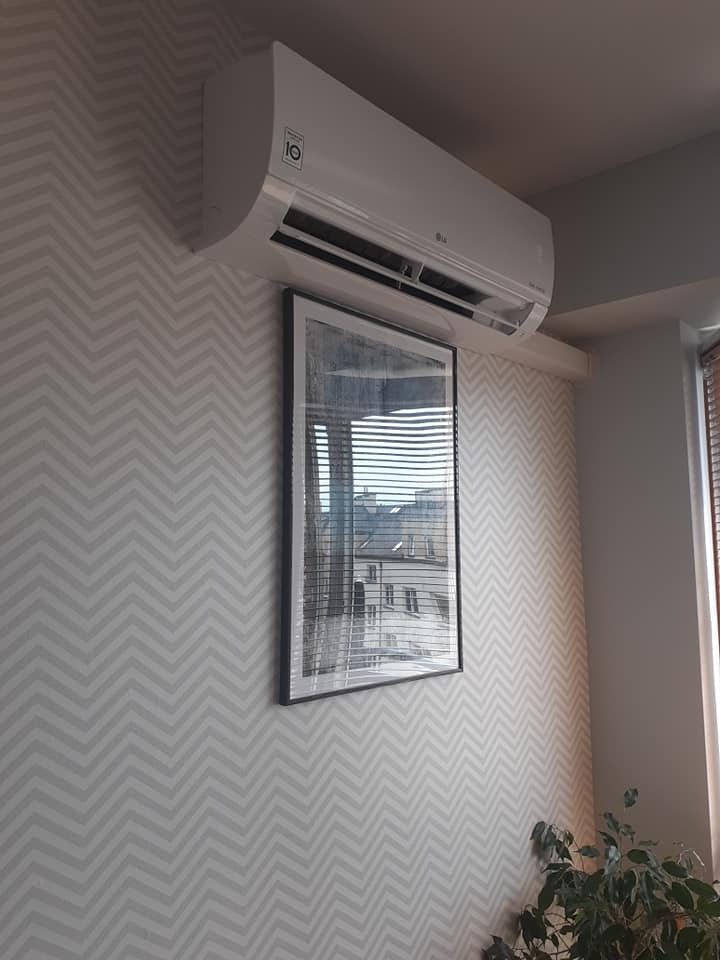 Biały klimatyzator LG zamontowany na ścianie z wzorem w zygzaki, pod nim wisi obraz z widokiem na budynki przez żaluzje.