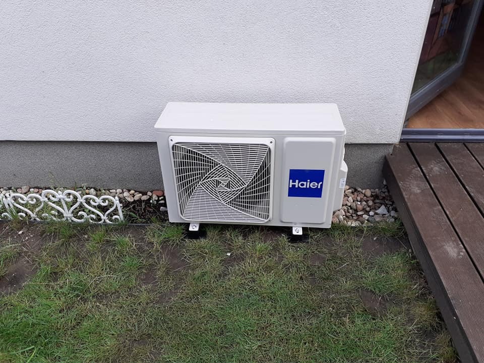Zewnętrzna jednostka klimatyzacji Haier zamontowana przy ścianie domu, widoczne trawnik, biały płotek ozdobny i fragment drewnianego tarasu.