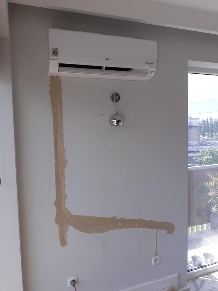 Biały klimatyzator LG na szarej ścianie w trakcie montażu, widoczne ślady po przewodach zakryte gipsem, okno z widokiem na zewnątrz.