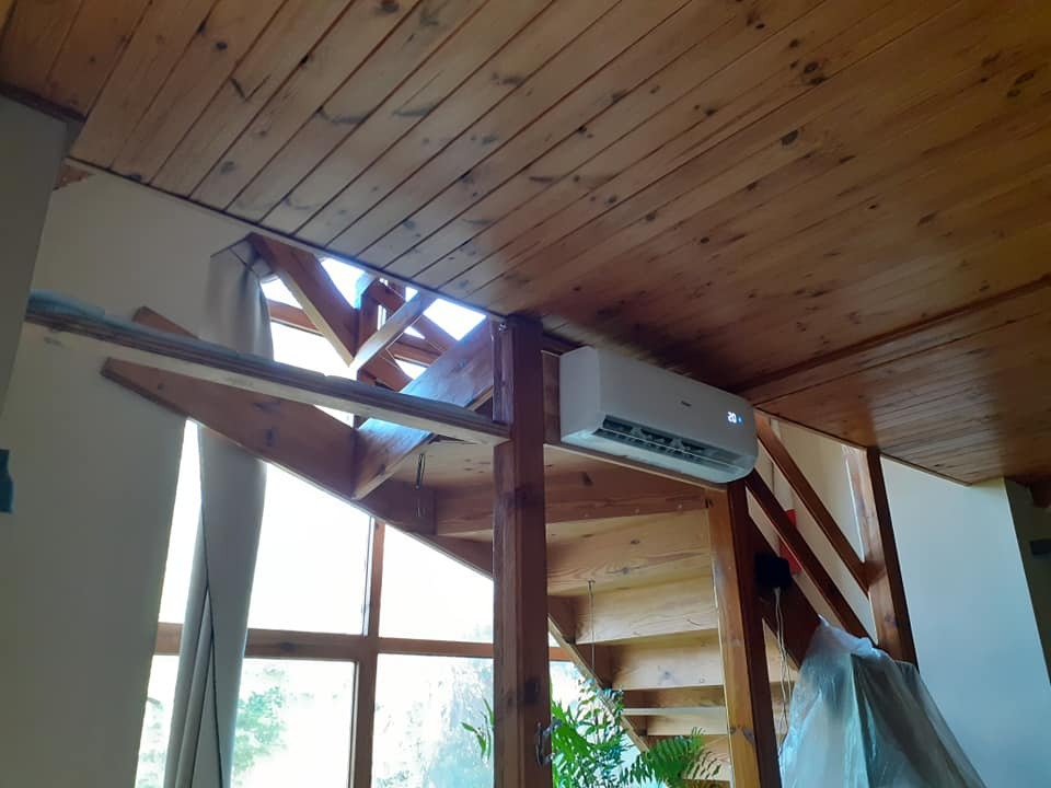 Klimatyzator zamontowany w domu z drewnianym stropem i schodami, widoczny wyświetlacz temperatury 20 stopni Celsjusza, instalacja w trakcie prac wykończeniowych.