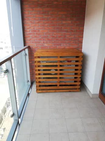 Klimatyzator na balkonie w drewnianej obudowie z listew, na tle ściany z czerwonej cegły.