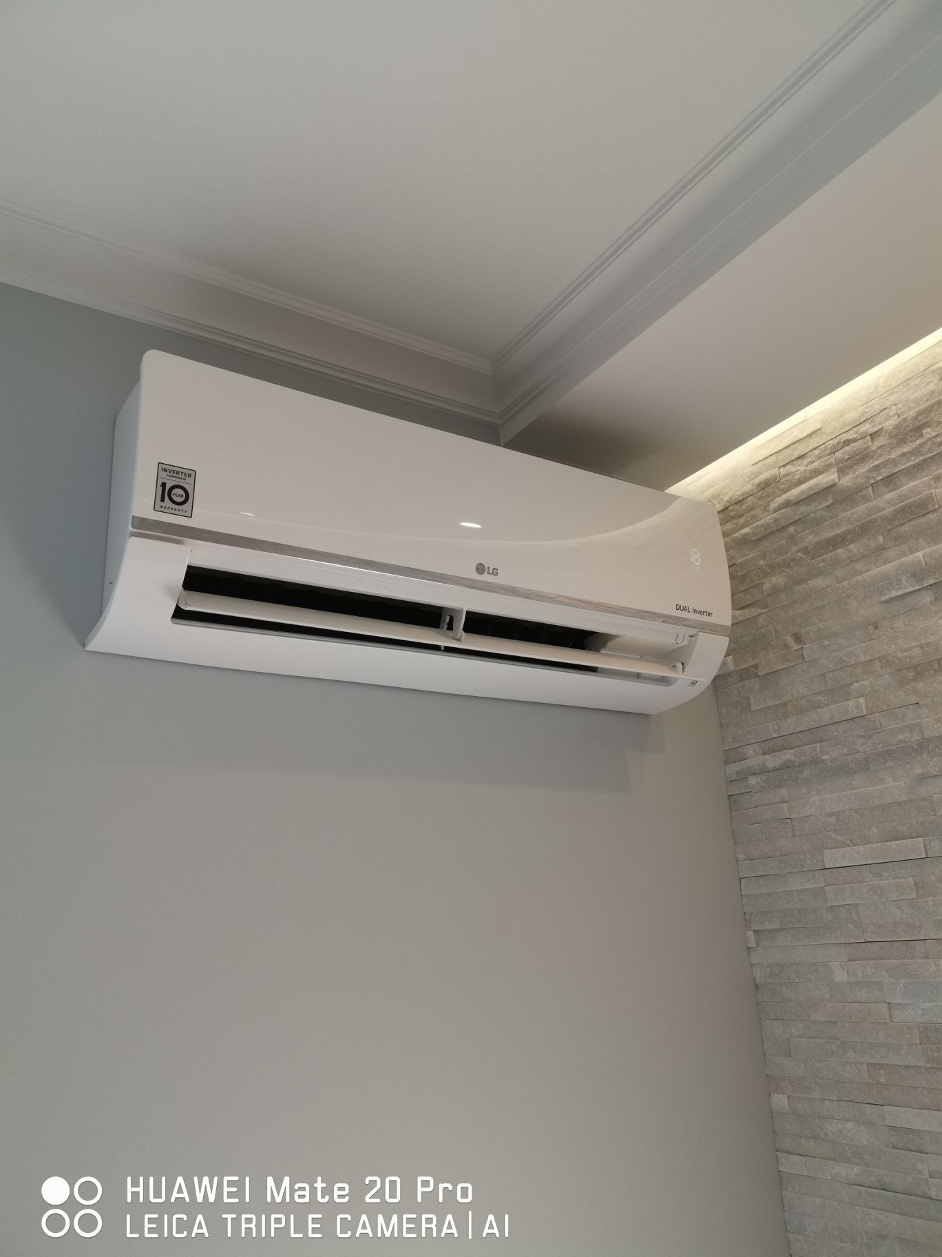 Biała klimatyzacja LG Dual Inverter zamontowana na szarej ścianie w pomieszczeniu z dekoracyjną ścianą z kamienia i oświetleniem LED.