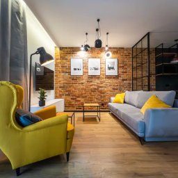 Odnova Studio Design AGNIESZKA FRYCZKE - Nowoczesny salon z ceglaną ścianą, szarą sofą z żółtymi poduszkami i żółtym fotelem typu uszak, drewniana podłoga, czarne lampy wiszące i grafiki na ścianie.