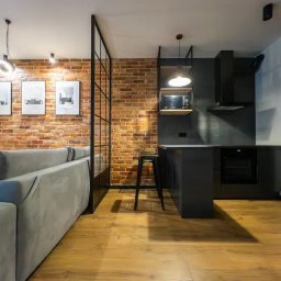 Odnova Studio Design AGNIESZKA FRYCZKE - Loftowe wnętrze: ceglana ściana, czarna kuchnia z wyspą i drewniana podłoga, oddzielone szklaną ścianką działową od salonu z szarą sofą i minimalistycznymi grafikami.