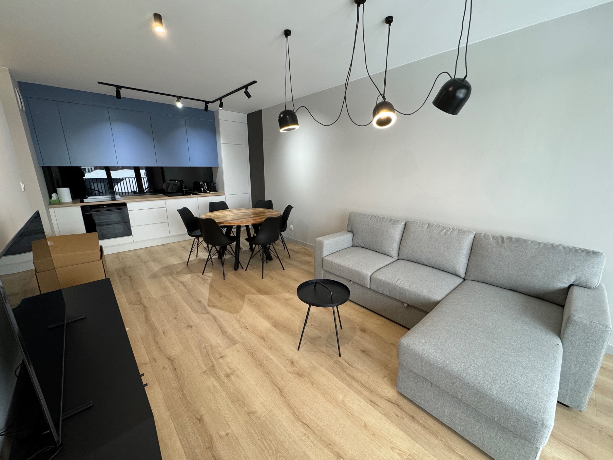 Jasne wnętrze mieszkania z drewnianą podłogą, szarą sofą narożną, okrągłym stołem z czarnymi krzesłami i niebieską zabudową kuchenną z czarnym szkłem na ścianie.