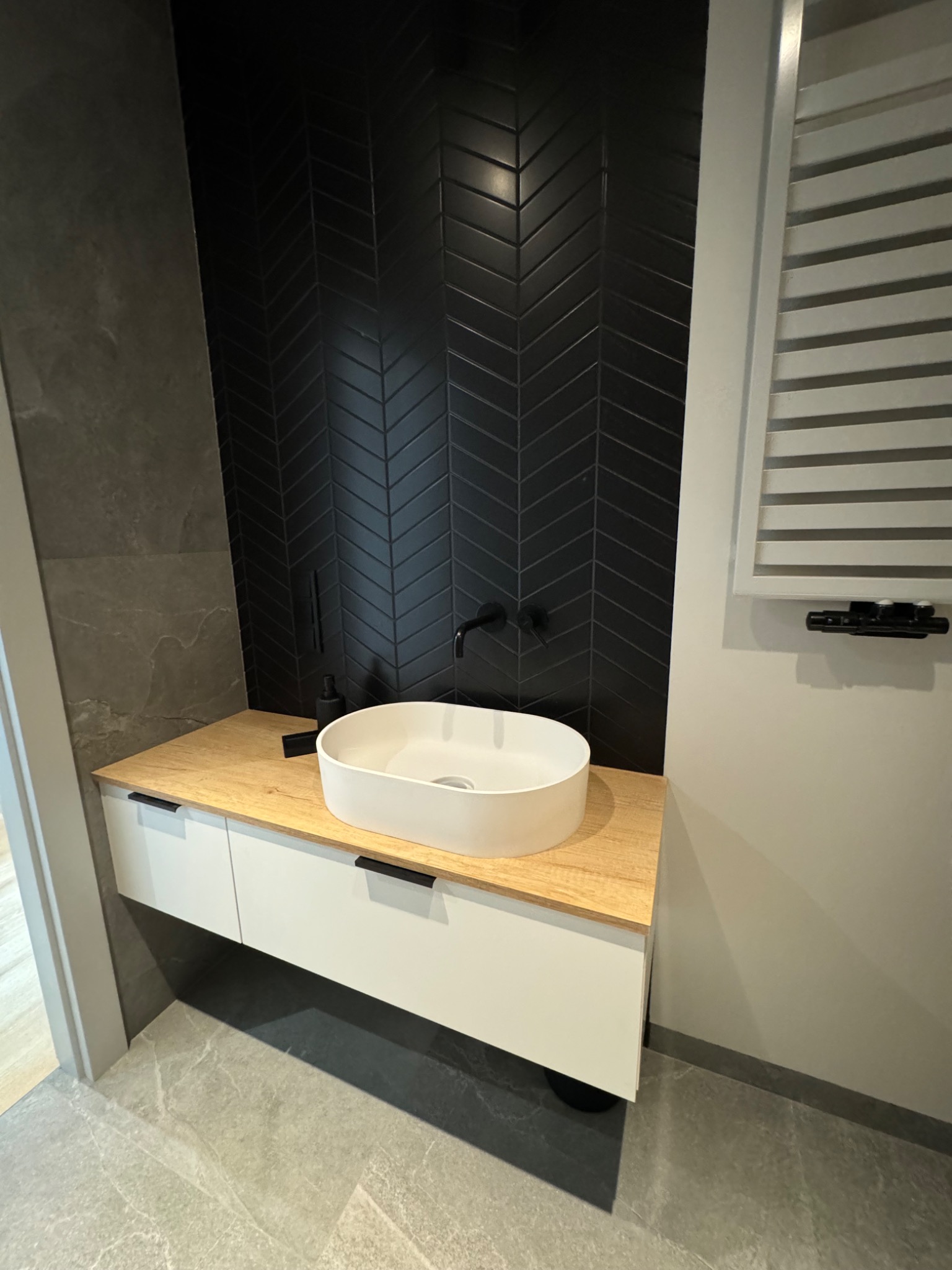 Nowoczesna łazienka z czarnymi płytkami ułożonymi w jodełkę na ścianie, owalną białą umywalką nablatową i drewnopodobnym blatem na białej szafce, szara marmurowa podłoga.