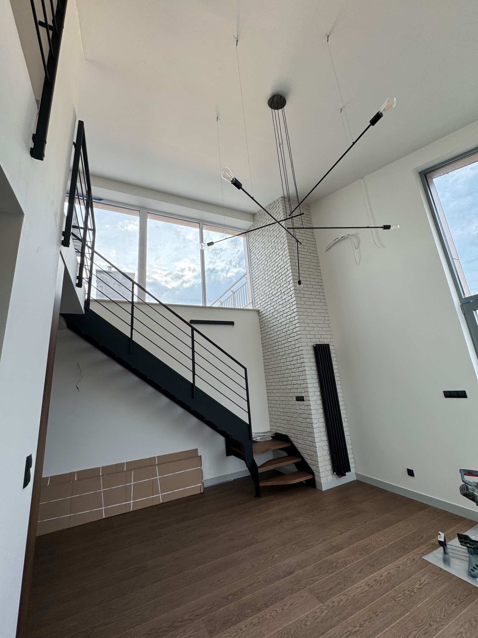 Wnętrze z czarnymi schodami z metalową balustradą, ceglaną ścianą pomalowaną na biało, nowoczesnym czarnym żyrandolem i drewnianą podłogą, widoczne kartony na podłodze i narzędzia sugerujące prace...