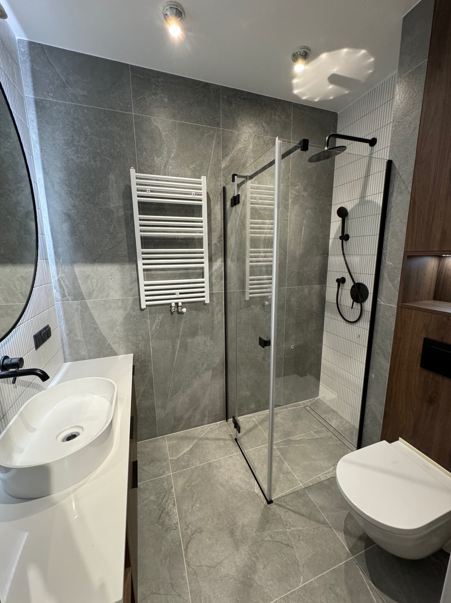 Nowoczesna łazienka z szarymi płytkami imitującymi kamień, kabiną prysznicową typu walk-in z czarnymi detalami, białą umywalką nablatową i grzejnikiem drabinkowym.