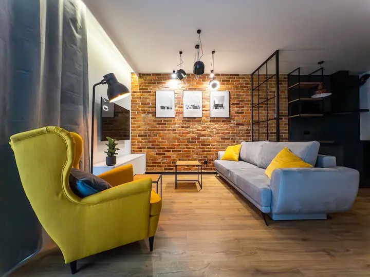 Nowoczesny salon z ceglaną ścianą, szarą sofą z żółtymi poduszkami i żółtym fotelem typu uszak, drewniana podłoga, czarne lampy wiszące i grafiki na ścianie.