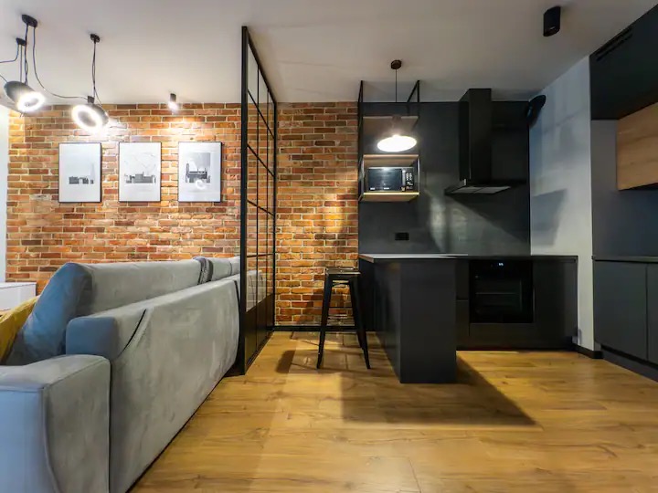 Loftowe wnętrze: ceglana ściana, czarna kuchnia z wyspą i drewniana podłoga, oddzielone szklaną ścianką działową od salonu z szarą sofą i minimalistycznymi grafikami.