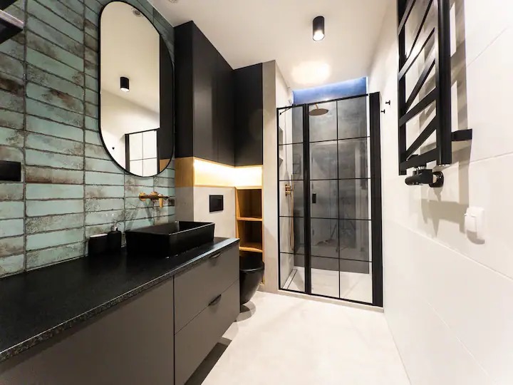 Nowoczesna łazienka z turkusowymi płytkami imitującymi cegłę, czarną armaturą i kabiną prysznicową typu walk-in z czarnymi szprosami. Wiszące szafki w kolorze grafitowym, podświetlane od dołu.