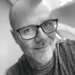 Czarno-biały portret uśmiechniętego mężczyzny w okularach, z krótką brodą i łysiejącą głową, skierowanego lekko w prawo, z miękkim oświetleniem.