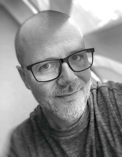 Czarno-biały portret uśmiechniętego mężczyzny w okularach, z krótką brodą i łysiejącą głową, skierowanego lekko w prawo, z miękkim oświetleniem.