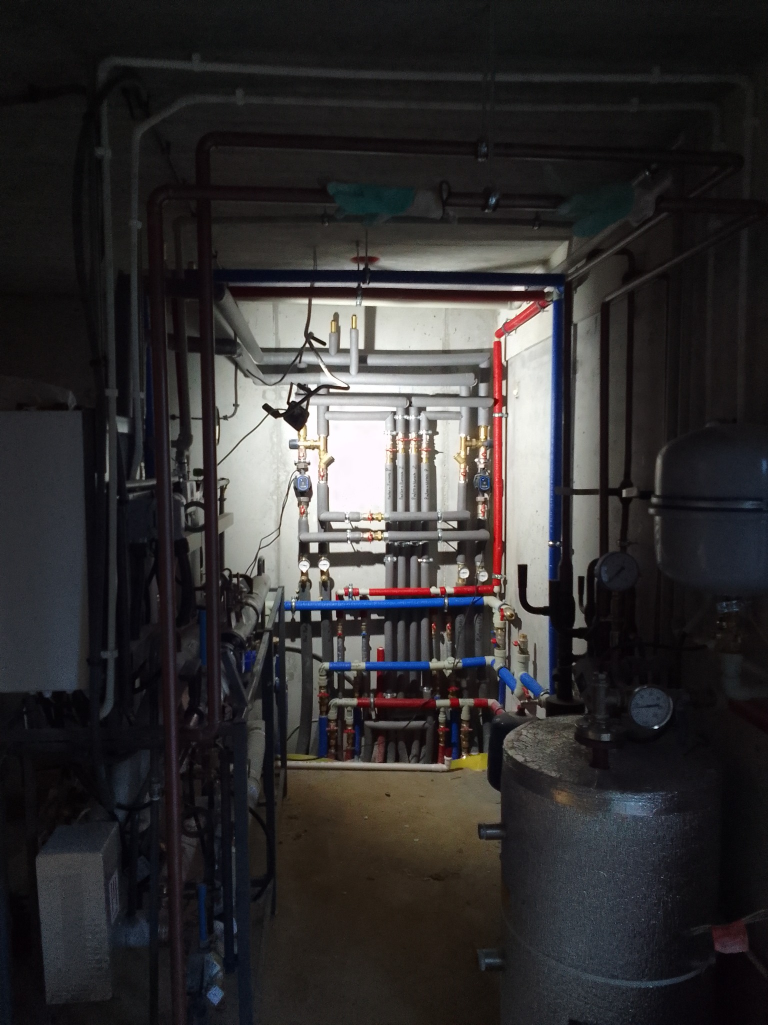 Węzeł cieplny w piwnicy z licznymi rurami, zaworami i manometrami, oświetlony punktowo, widok z perspektywy podłogi w kierunku ściany z instalacjami.